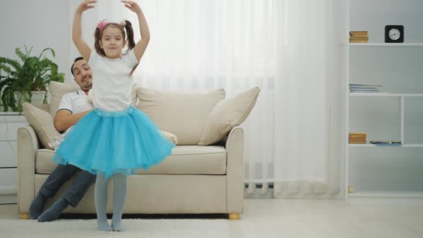 Das kleine Mädchen dreht sich um wie eine Ballerina. Vater sitzt auf dem Sofa, schaut sie lächelnd an. — Stockvideo