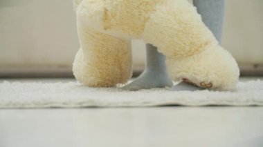 Kırpılmış küçük kız bacakları beyaz halıda duruyor, kolyesinde pelüş bir ayı tutuyor, patisini ayaklarına koyuyor ve ona nasıl dans edileceğini öğretiyor..