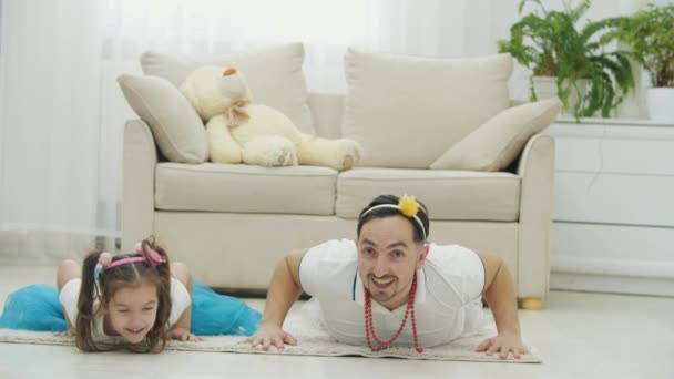 Lustig far och dotter bär fina huvudbonader, går armhävningar, på mattan. — Stockvideo