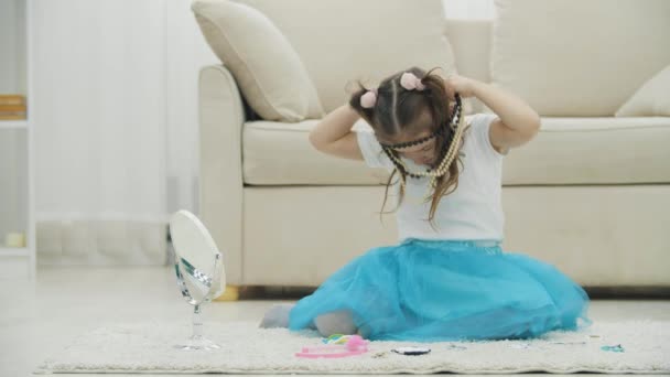 Nettes kleines Mädchen mit Zöpfen sitzt auf dem Teppich und trägt verschiedene Halsketten und Armbänder. — Stockvideo