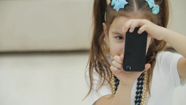 Обрезанное видео милой маленькой девочки с косичками, носящей ожерелье и головной убор, держащей черный телефон, фотографирующей. — стоковое видео