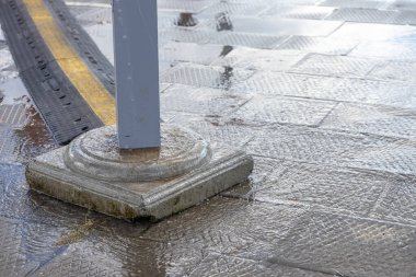 Beton bir kutup tabanını çevreleyen parıldayan su yansımaları ve şehir yüzeyi koşullarını gösteren siyah-sarı kablo koruyucusu ile ıslak desenli gri asfalt.