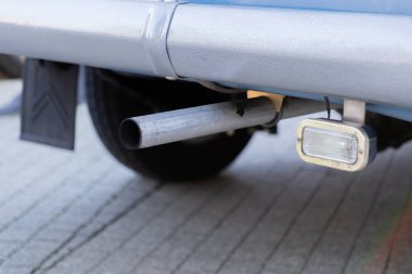 Yakın çekim, yıpranmış metalik egzoz sistemini ve eski bir aracın dikdörtgen arka lambasını gösteriyor. Bu da düzenli bakım ve araç emisyonlarına duyulan ihtiyacı gösteriyor.