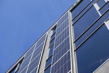 Düşük açılı görüntü, yenilenebilir enerji çözümlerini temsil eden, karanlık pencereleri ve güneş fotovoltaik panelleri olan çağdaş bir ofis binasının dış görüntüsünü yakalıyor.