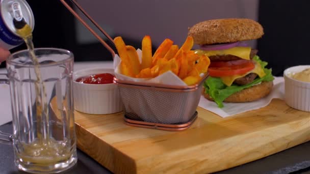 Verter cerveza en una taza y una hamburguesa americana jugosa con papas fritas crujientes — Vídeo de stock