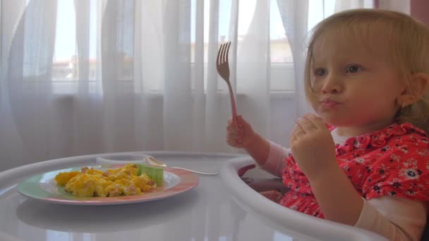 Uma menina bonita comendo ovos mexidos em uma cadeira de bebê — Vídeo de Stock