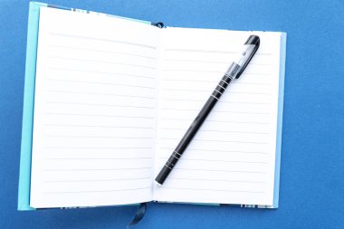 Mavi arka planda açık bir defter var. Siyah bir peni var.