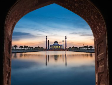 Beautiful mosque reflected in tranquil water at sunset, viewed through a decorative archway, creating a serene and spiritual atmosphere