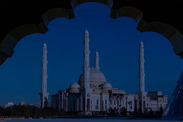 Beautiful grand mosque with multiple tall minarets and domes framed by an arch, captured during evening blue hour under clear sky.