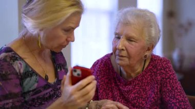 Orta yaşlı kızıyla aynı masada oturan mutlu, gülen yaşlı bir kadın. Ailesini ya da telefonda seyahat fotoğraflarını izliyor. Gülümseyen sarışın kadın. Yaşlı dinleyen, akıllı telefon kullanan..