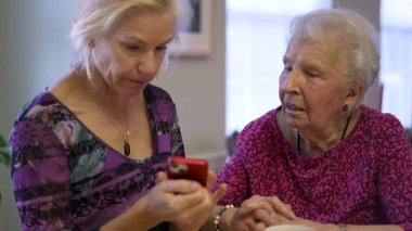 Yetişkin kızıyla masada oturan mutlu, orta yaşlı bir kadın. Ailesini ya da telefonda seyahat fotoğraflarını izliyor. Gülümseyen sarışın kadın. Yaşlı dinleyen, akıllı telefon kullanan..