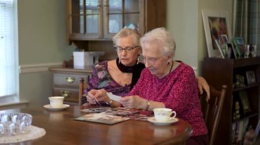 Yaşlı ve mutlu bir kadının yavaş çekimde yemek odasında kızıyla fotoğraflara bakması. Aileyle fotoğraflara bakıyorum. Beyin eğitimi. Bellek etkinliği.