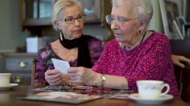 Yaşlı ve mutlu bir kadının yavaş çekimde yemek odasında kızıyla fotoğraflarına bakması. Aileyle fotoğraflara bakıyorum. Beyin eğitimi. Bellek etkinliği.
