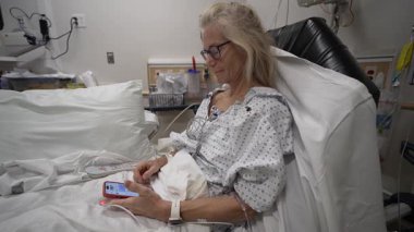 Yetişkin bir kadın hastane yatağında, önlük giymiş ve telefonunu bağlı kalmak için kullanıyor. Acil servis onun dayanıklılığını ön plana çıkarıyor..