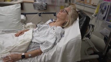 Winchester, VA, ABD - 09 03 2025: Olgun bir kadın hastane yatağında dinleniyor ve acil serviste tıbbi yardım alıyor. Personel destek sağlarken, monitörler hayati belirtileri gösterir.