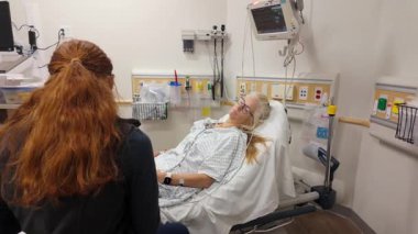 Winchester, VA, ABD - 09 03 2025: Olgun bir kadın hastane yatağında dinlenirken, arkadaşı duygusal destek sağlıyor. Oda tıbbi cihazlar ve temel bakım malzemeleriyle donatılmıştır..