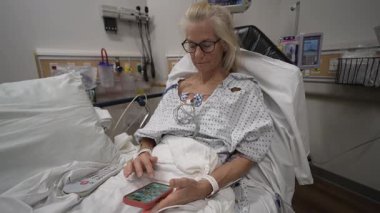 Winchester, VA, ABD - 09 03 2025: Olgun bir kadın hastane acil servisinde tıbbi önlük giymiş ve tıbbi personelden tedavi ve bakım görürken mobil cihazını kullanıyor.