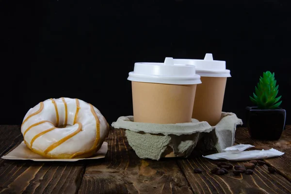 Paper cups of coffee, sugar in bags, donuts. Cactus. Wooden table. Black background.