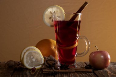 Bir bardak portakal kabuklu sıcak şarap ve bir dilim limon, tarçın çubukları, yıldız anasonu ve turuncu arka plandaki ahşap bir masada doğranmış narenciye ve elma..