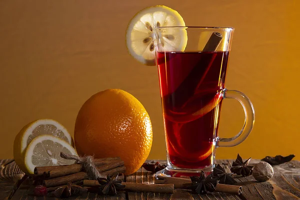 Glass of mulled wine with orange peel and a slice of lemon, cinnamon sticks, star anise and chopped citrus on a wooden table on an orange background. — Stock Photo, Image