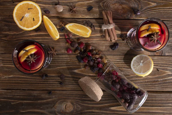 Horní pohled na dvě sklenice se svařeným vínem s pomerančovou slupkou a plátkem citronu, skořicovými tyčinkami, sklenicí sušeného ovoce na dřevěném stole. — Stock fotografie