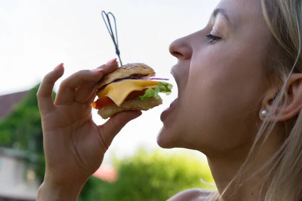 Woman eating sandwich. She opened her mouth, holding a hamburger on his outstretched hands and closed her eyes. — Stock Photo, Image