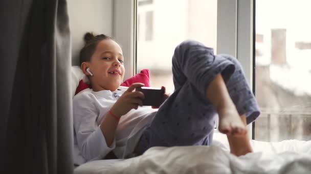 Çocuk yakınındaki pencere pervazına üzerinde oturan bir akıllı telefon kullanan ve kablosuz kulaklıklar müzik dinler — Stok video
