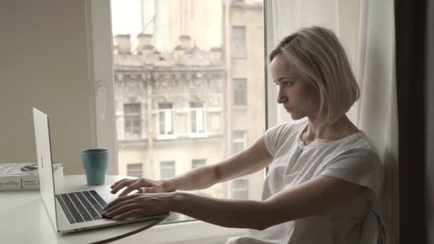 Młoda kobieta siedzi w pobliżu okna i w domu za pomocą komputera przenośnego — Wideo stockowe