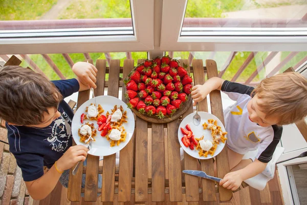 Due ragazzi stanno mangiando waffle con fragole e gelato su un tavolo di legno sul balcone — Foto Stock