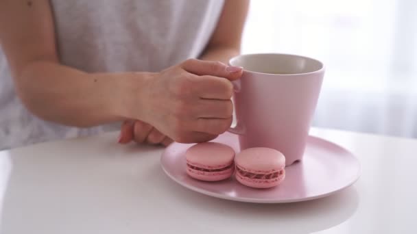 Молодая белая женщина пьет чай с розовым макароном на белой кухне — стоковое видео