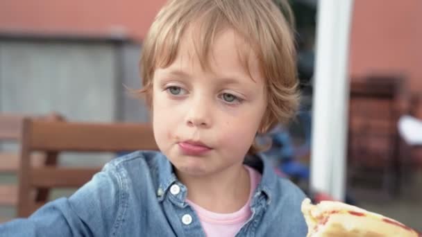 Menino de 5 anos em camisa jeans comendo delicioso cachorro quente . — Vídeo de Stock