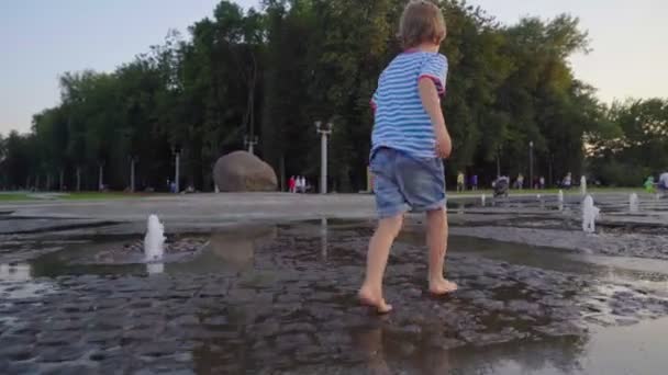 Un niño pequeño en pantalones cortos de mezclilla y zapatos amarillos corre en la fuente en la noche de verano — Vídeo de stock