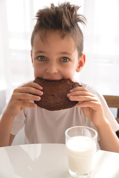 Cute boy sitting on kitchen eating chocolate cookie with milk — Stock Photo, Image