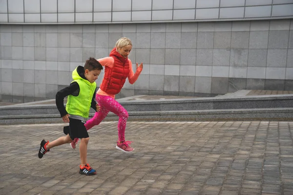 Die sportliche junge Frau und der Junge laufen vor dem Hintergrund der urbanen Landschaft. Familiensport. — Stockfoto