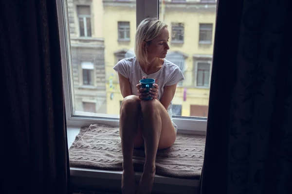 Menina calma com xícara de chá ou café sentado e bebendo no peitoril da janela em casa . — Fotografia de Stock