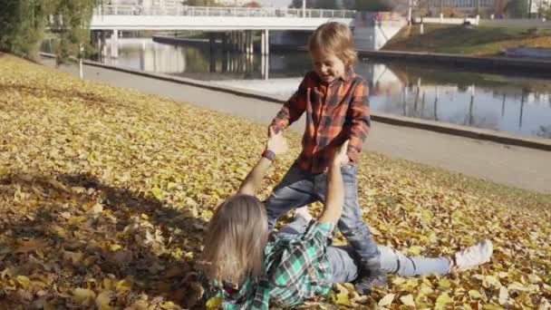 Мать и сын веселятся в осеннем парке и лежат в куче желтых листьев. Мама обнимает мальчика — стоковое видео