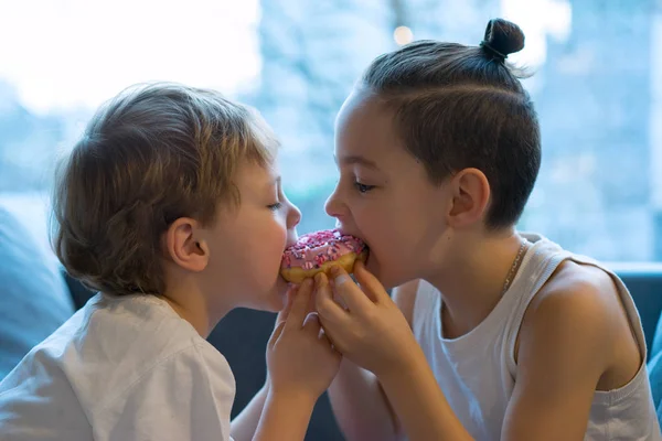 Two kids bite off a donut and having fun. two boys together bite from the donut. children enjoy a donut with strawberry frosting. divide the a donut in half. — Stock Photo, Image