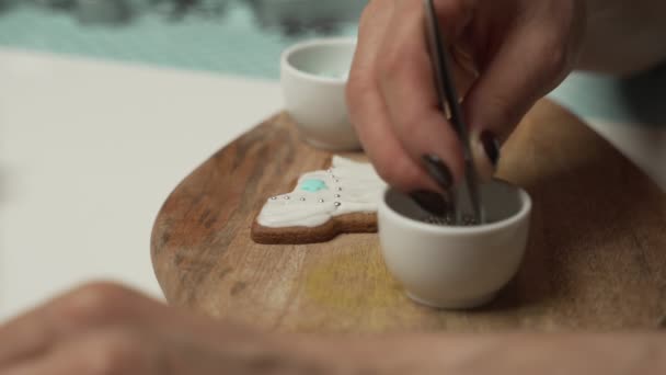 Una chica joven decora galletas de jengibre noche de invierno de Navidad. La mujer dibuja hielo en la casa de miel de jengibre. Mesa marrón madera. espacio de copia. Galleta en blanco casa de jengibre, listo para decorar . — Vídeos de Stock