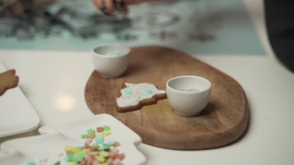 Una chica joven decora galletas de jengibre noche de invierno de Navidad. La mujer dibuja hielo en la casa de miel de jengibre. Mesa marrón madera. espacio de copia. Galleta en blanco casa de jengibre, listo para decorar . — Vídeos de Stock