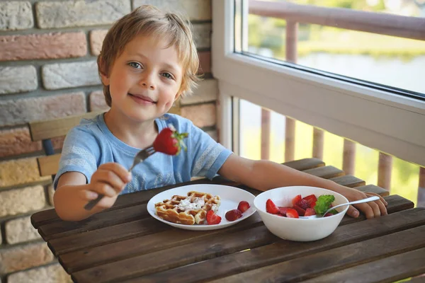 Счастливый 5-летний кавказский мальчик ест на завтрак венские вафли с мороженым и клубникой — стоковое фото