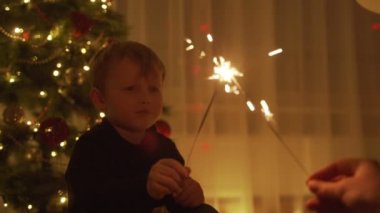 Çocuk tutan bir maytap ve gülüyor. Havai fişekleri. Havai fişekleri onun elinde. Yeni yıl ruh. Noel ağacı. Işıklar. Yakışıklı çocuk. Çocuk.