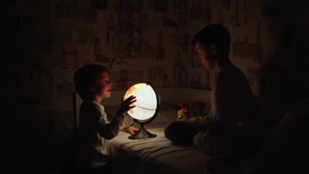 Dos chicos sentados en la cama por la noche girando el globo y sueña con viajar . — Vídeos de Stock