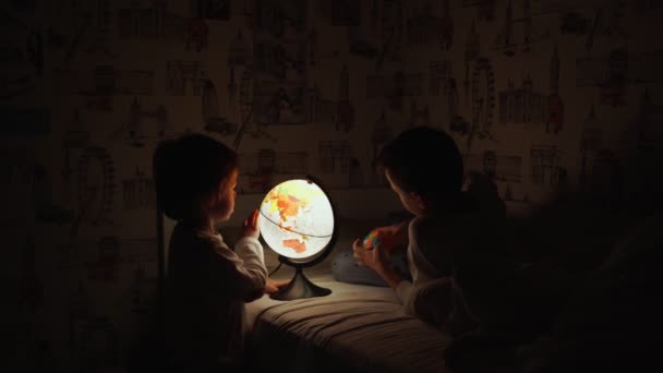 Dos chicos sentados en la cama por la noche girando el globo y sueña con viajar . — Vídeos de Stock