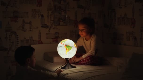 Twee jongens zitten in bed bij nacht draaien de globe and dromen van reizen. — Stockvideo