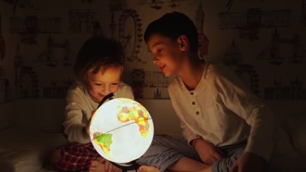 Dos chicos sentados en la cama por la noche girando el globo y sueña con viajar . — Vídeos de Stock