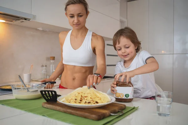 Молодая спортивная женщина с ребенком приготовления блинов на кухне — стоковое фото