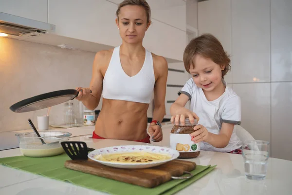 Молодая спортивная женщина с ребенком приготовления блинов на кухне — стоковое фото