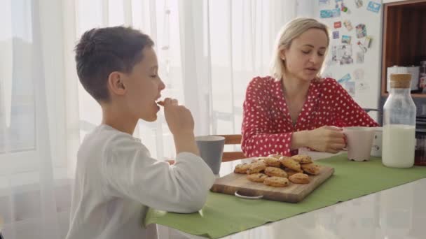 사랑 스러운 귀여운 백인 소년 금발 머리를 가진 그의 젊은 어머니 옆에 쿠키를 먹고 있다. — 비디오