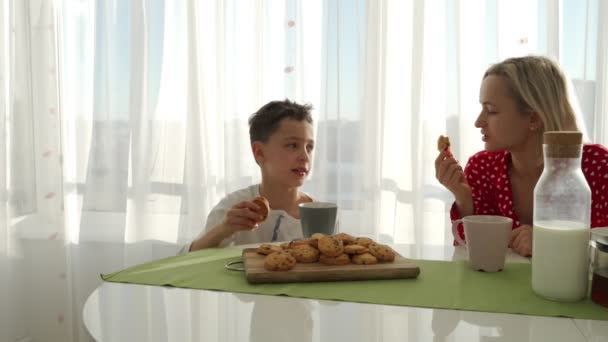 Un adorabile ragazzo caucasico carino sta mangiando biscotti accanto alla sua giovane madre con i capelli biondi. Il fratellino ruba biscotti. . — Video Stock