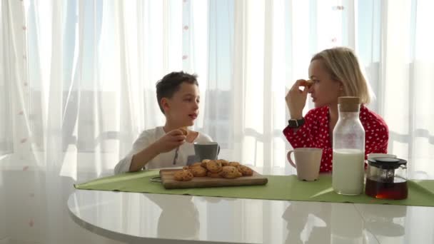 愛らしいのかわいい白人少年は、ブロンドの髪を持つ若い母親の横にクッキーを食べています。弟はクッキーを盗んでいます。. — ストック動画